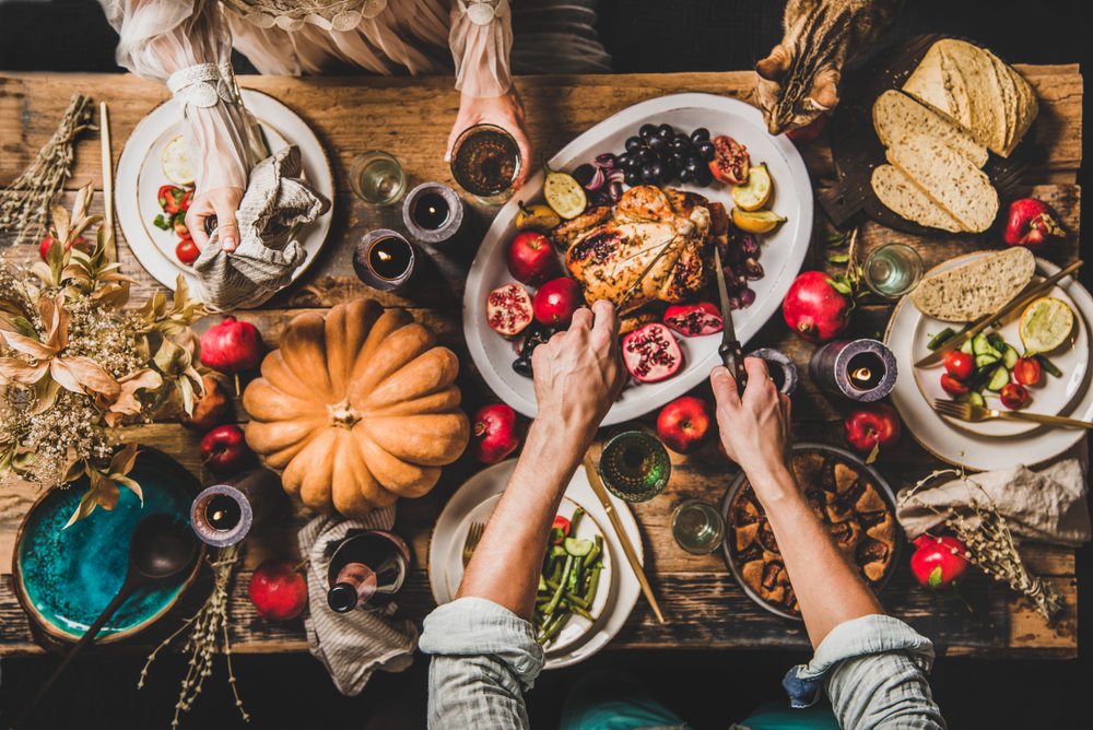 A top view of a rustic wooden table set for a festive meal. In the center is a roasted poultry dish surrounded by various fruits, bread, and vegetables. Multiple hands are seen serving food and reaching for drinks, with a large pumpkin and candles adding to the autumn ambiance, perfect for unwinding after a busy day at the vet clinic.